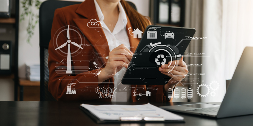 Image of a woman from neck down in a brown jacket and white shirt holding a tablet while seated at a desk. A n open laptop and a clipboard lies on the desk before her. Superimposed across the image are graphs, charts and symbols indicating she is working on green technology ideas.